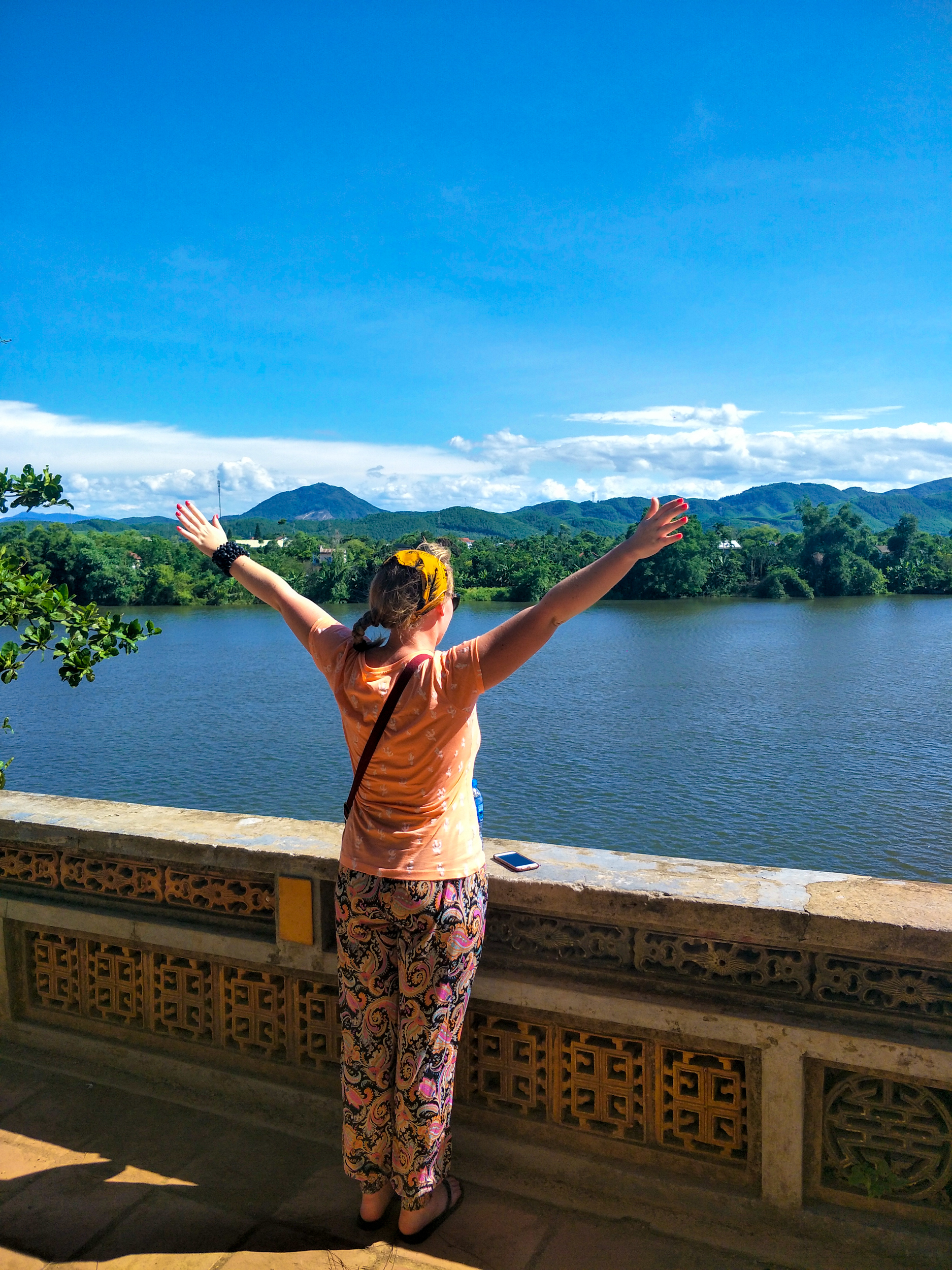 Woman with arms open wide overlooking a beautiful river and distant green mountains in Vietnam.