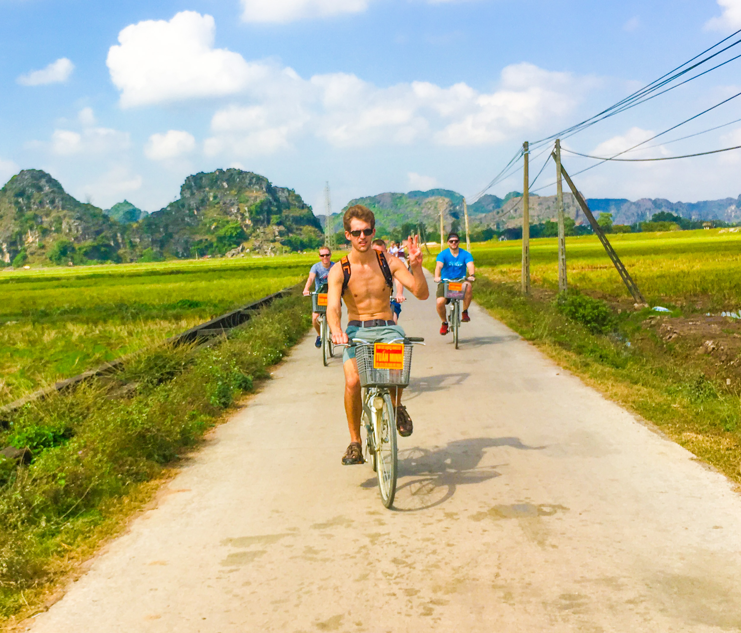 Three friends cycle on a sunny day through green rice paddies with dramatic limestone mountains in Vietnam.