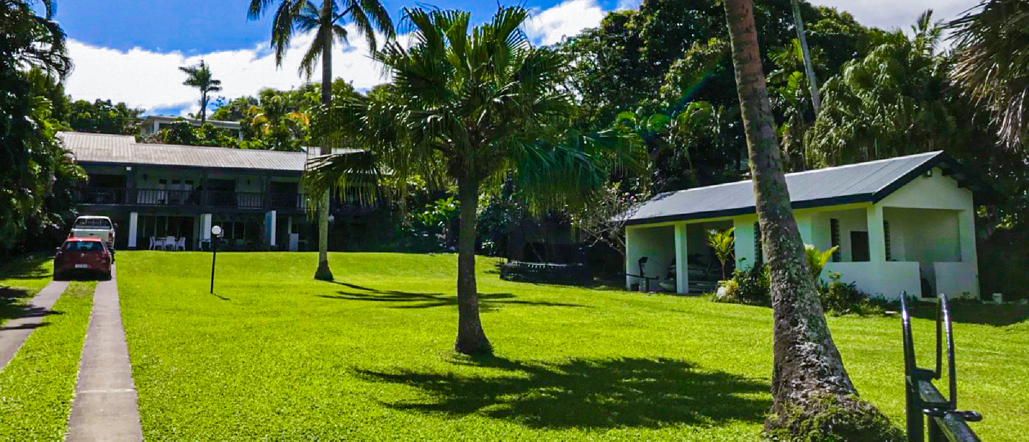 A lush green lawn with tall palm trees, two buildings, and a red car in tropical Fiji.
