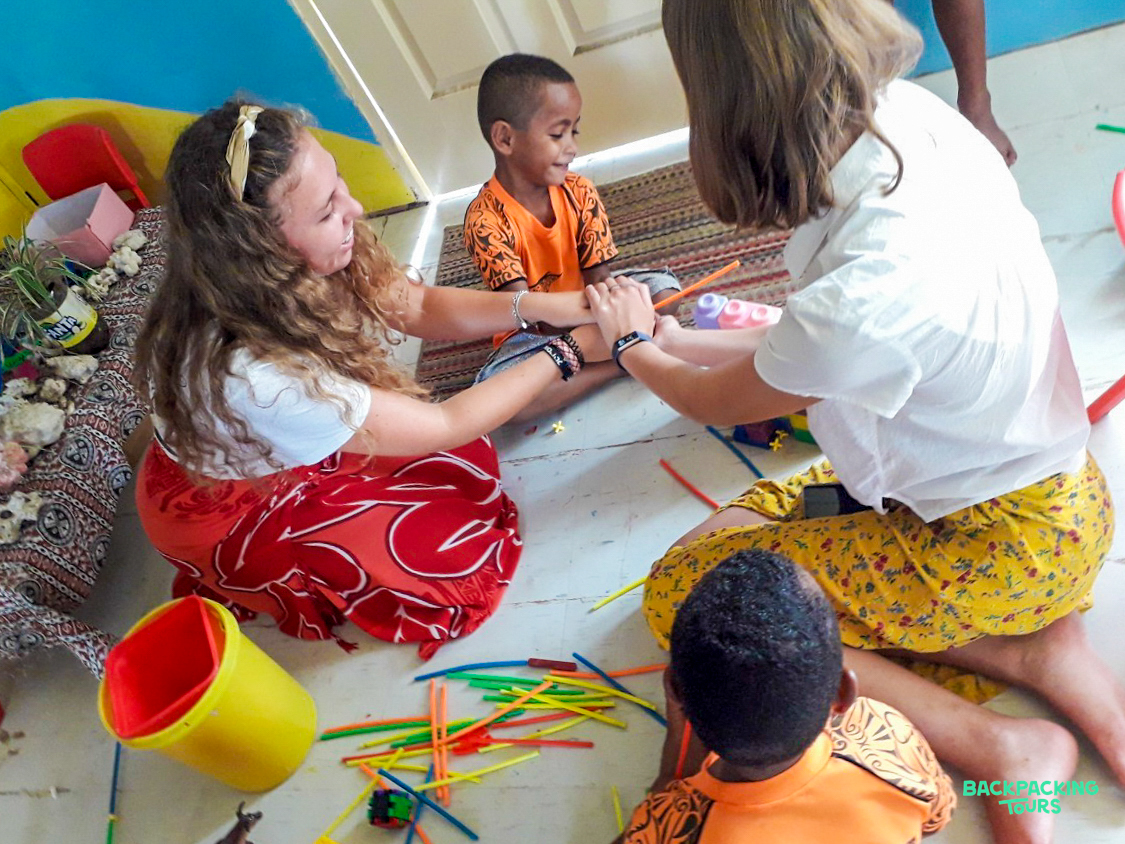Two young women and two children happily play with colorful sticks on the floor in a Fijian kindergarten.