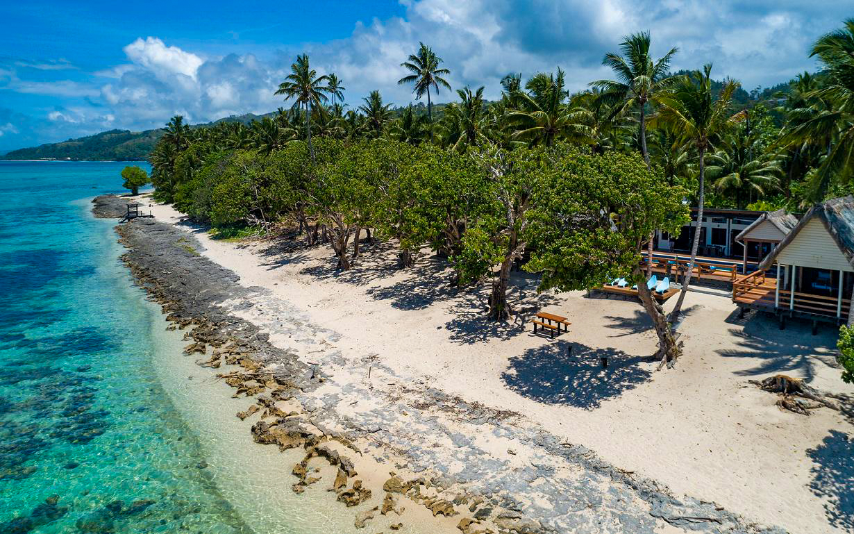 Tropical beach in Fiji with clear ocean, sandy shore, lush trees, and small bungalows.