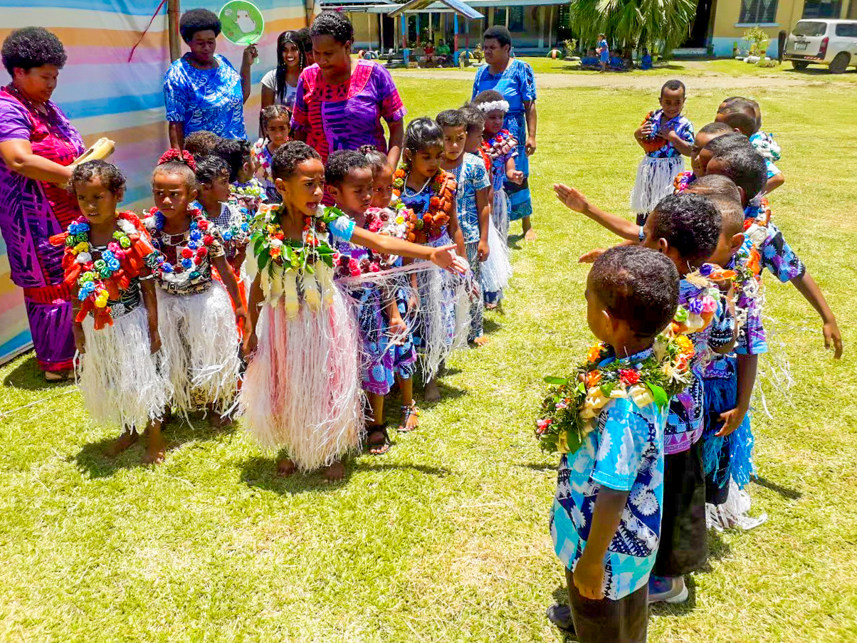 Young children in vibrant leis and traditional skirts celebrate a kindergarten graduation outdoors in Fiji.