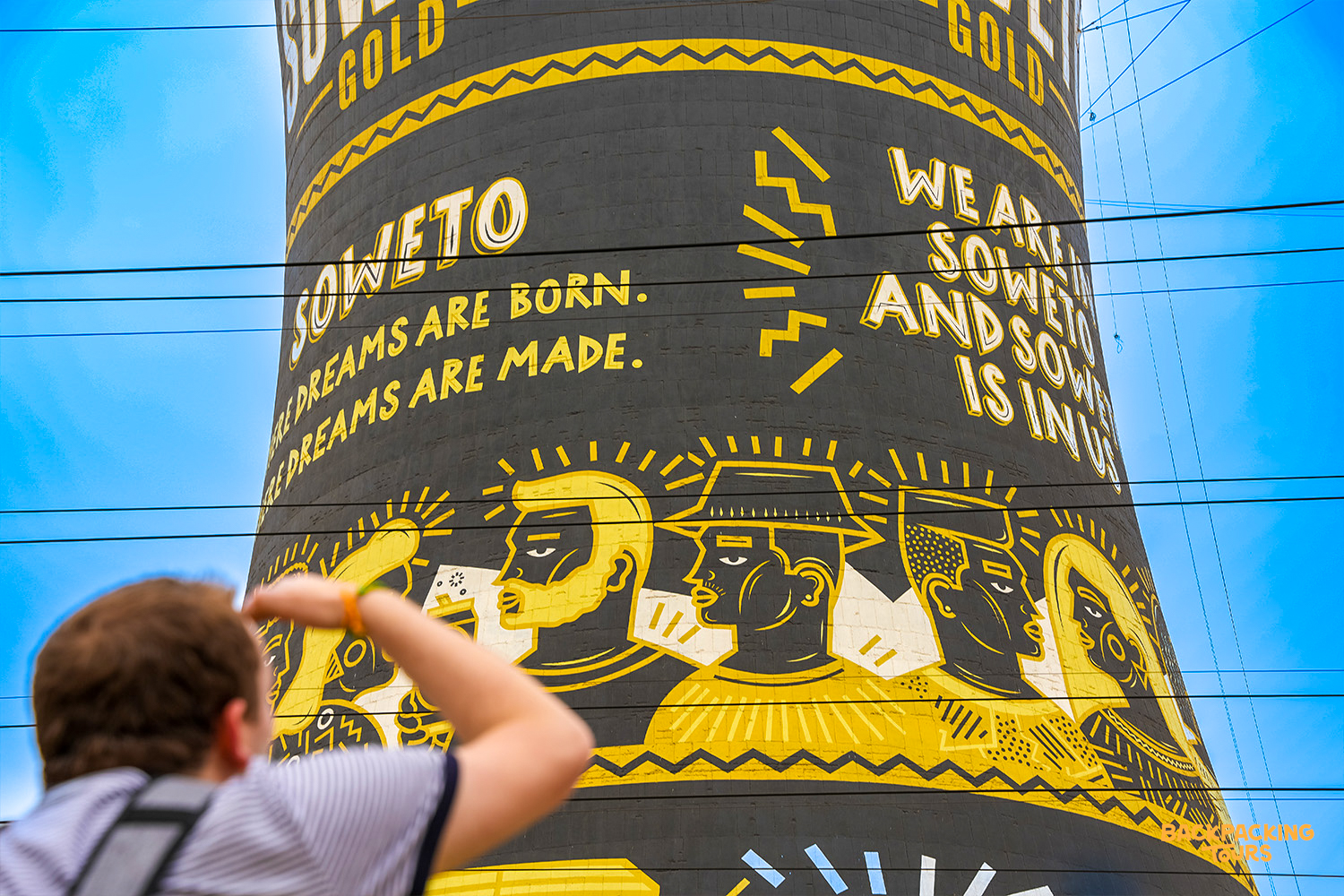 A person looks up at the vibrant Orlando Towers mural in Soweto, featuring inspiring text and portraits.