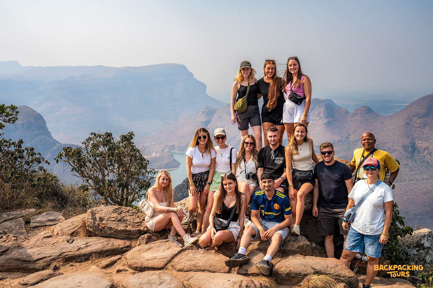 A group of happy backpackers posing on a rocky viewpoint overlooking the vast Blyde River Canyon.