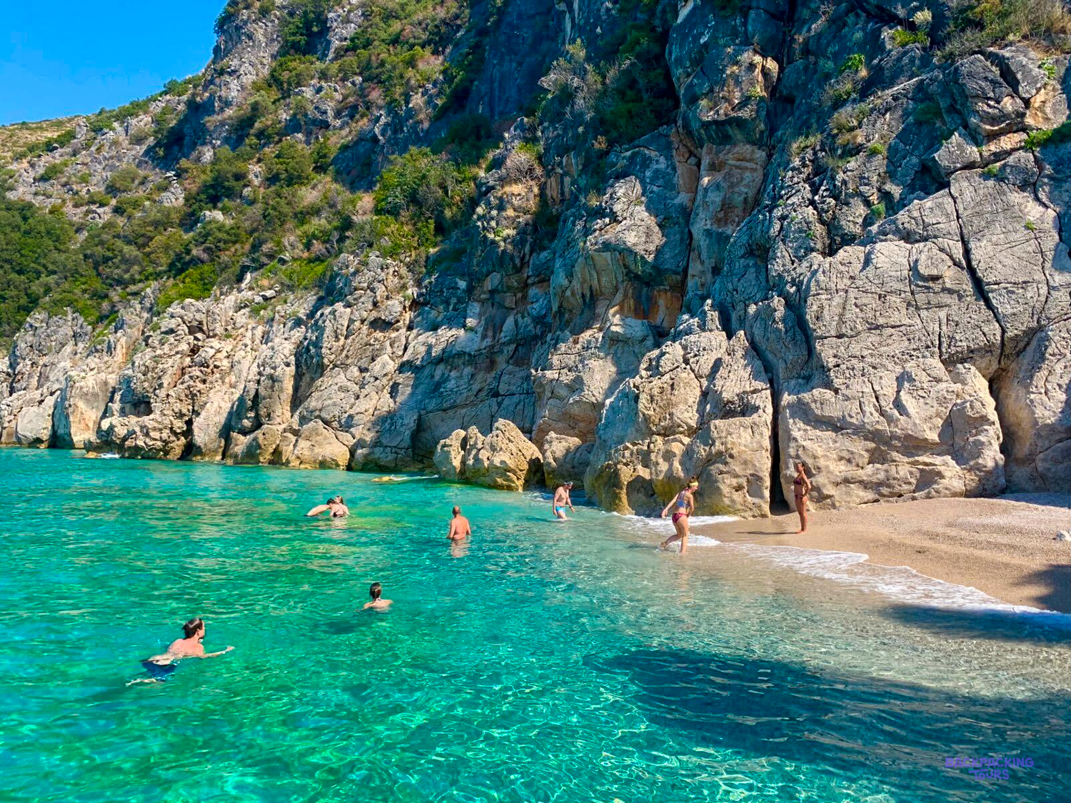 Swimmers in turquoise water below limestone cliffs at a pebble beach near Himarë