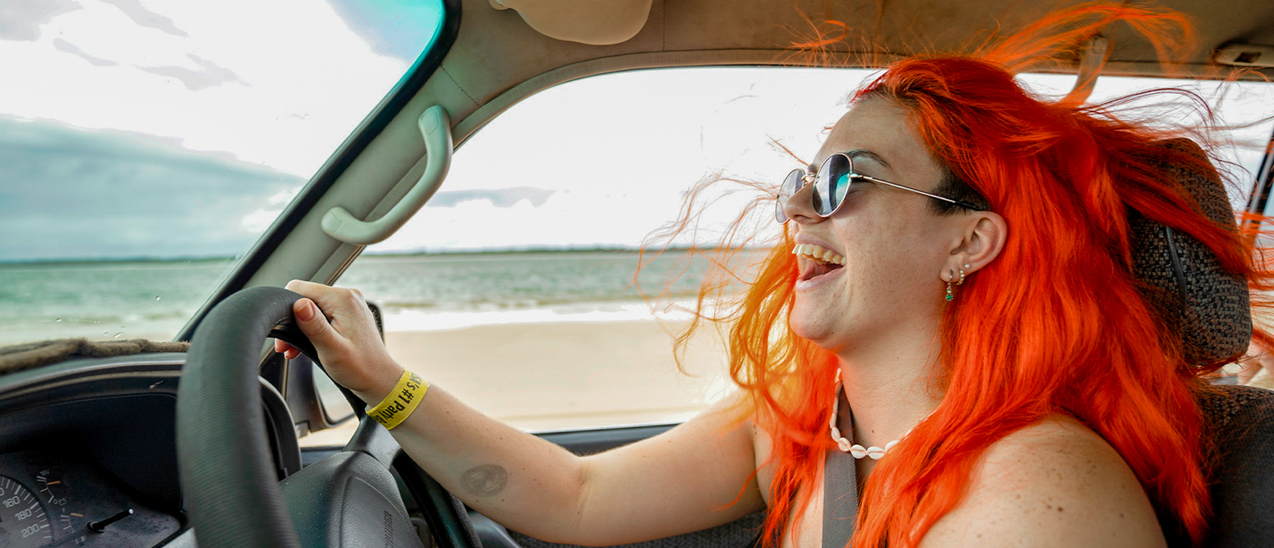 A joyful woman with vibrant orange hair drives a vehicle along Rainbow Beach.