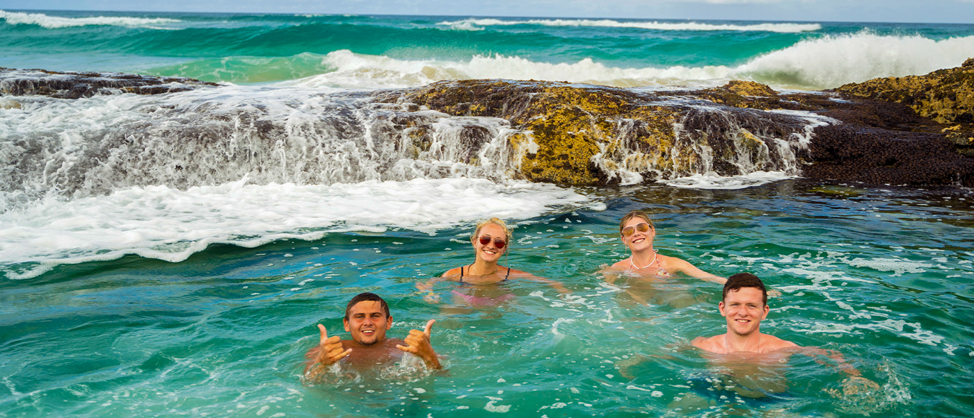 Happy backpackers swim in the clear Champagne Pools on K'gari, with waves breaking over rocks.