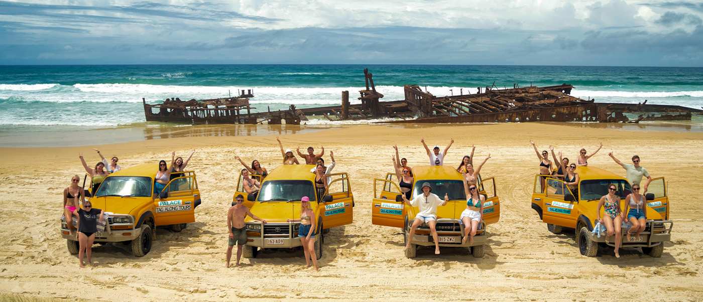 Happy backpackers pose with yellow 4WDs on Fraser Island beach, a historic shipwreck in the background.