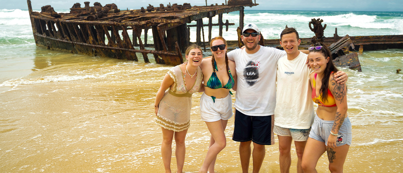 Five happy backpackers pose in front of the Maheno shipwreck on Fraser Island, Australia.