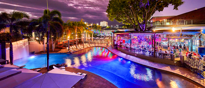 A vibrant night scene at a tropical resort in Cairns, featuring a large pool and lively bar.