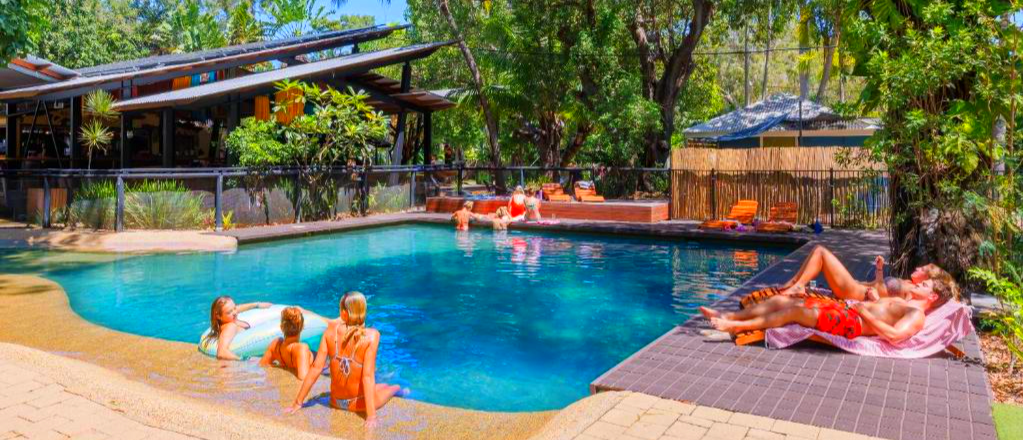 Travelers relax by a resort pool on Magnetic Island, sunbathing and swimming under green trees.