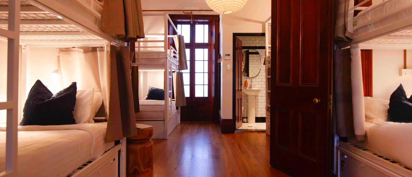 A warmly lit, clean hostel dorm room with white bunk beds and privacy curtains in Sydney.