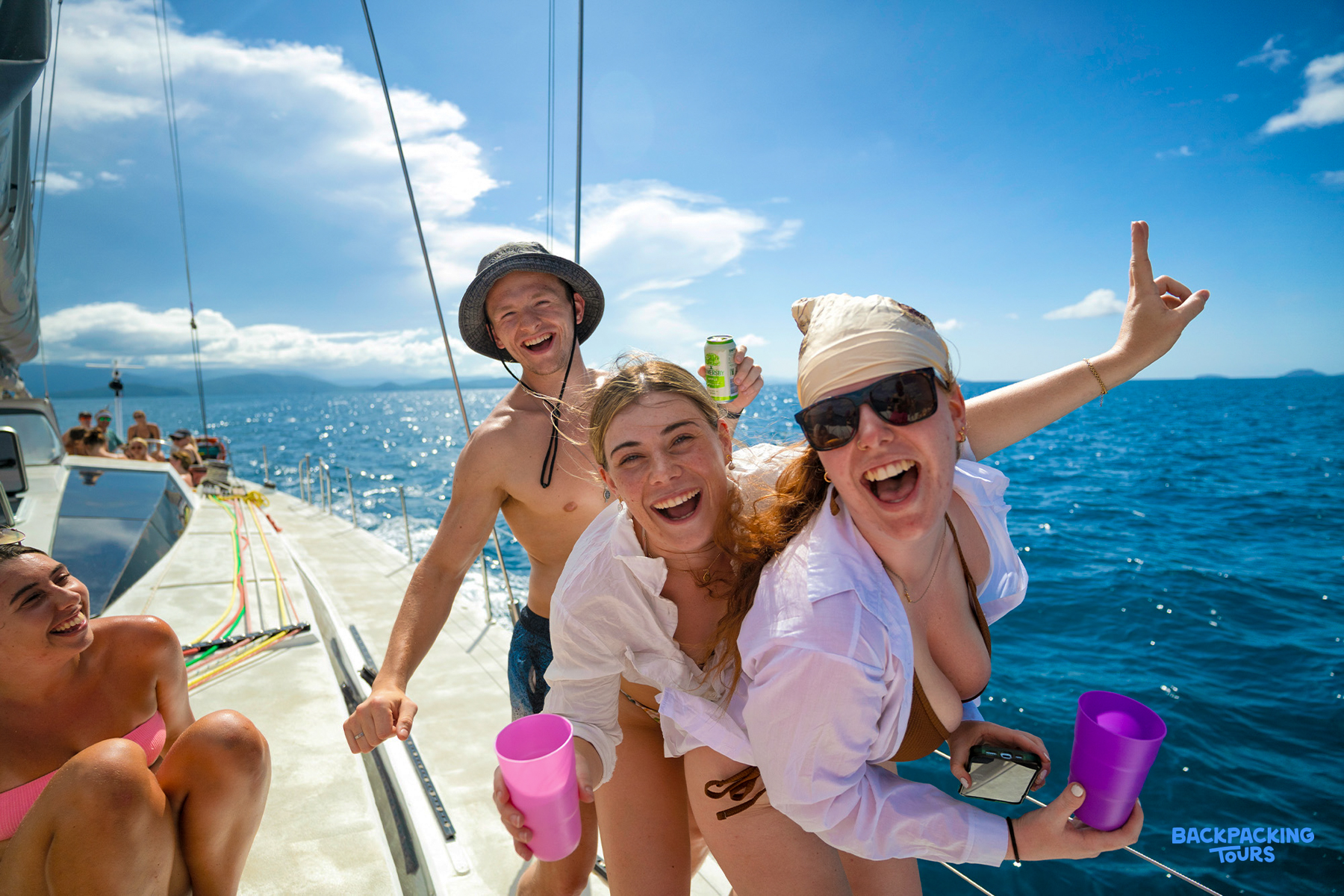 Three travellers laughing together on the deck of a sailing boat in the Whitsundays, drinks in hand, open blue sea behind them.