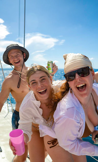 Three travellers laughing together on the deck of a sailing boat in the Whitsundays, drinks in hand, open blue sea behind them.
