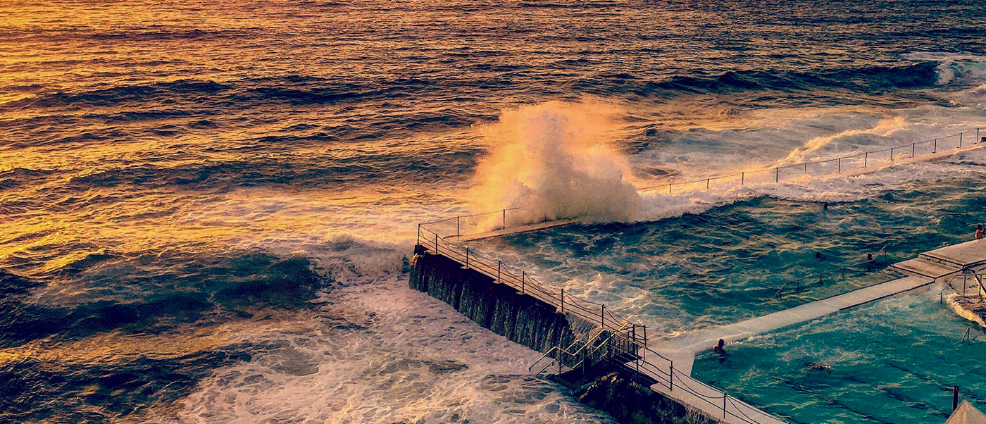 Waves crash dramatically over the iconic Bondi Icebergs ocean pool at sunset, with swimmers enjoying the water.