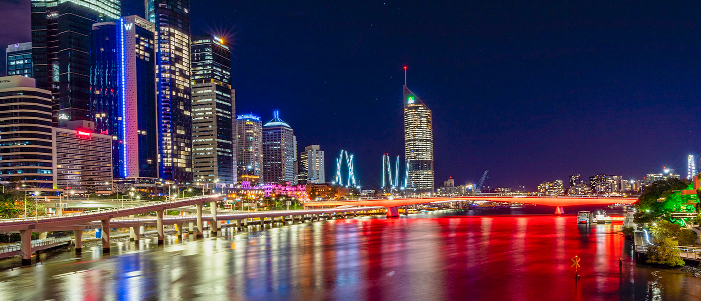 The vibrant Brisbane city skyline at night, with illuminated buildings reflecting colorful lights on the river.