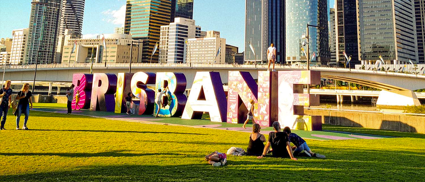 Visitors relax on the grass in front of the colorful Brisbane sign with a city skyline.