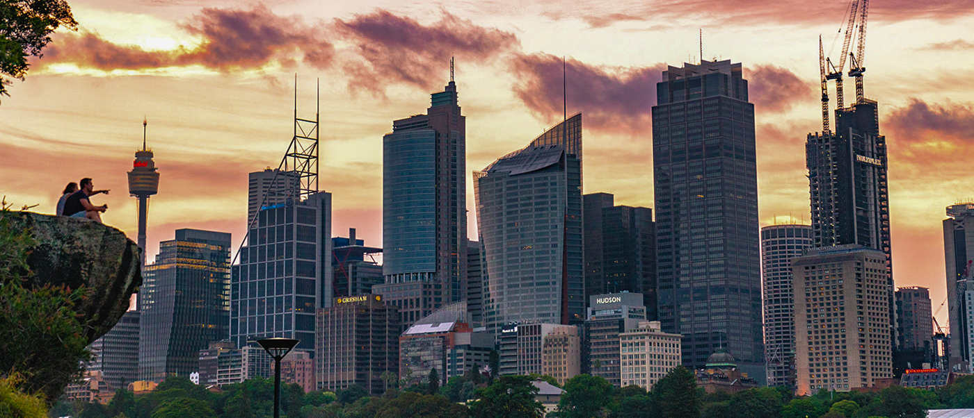 Two people sit on a rock, admiring the expansive Sydney city skyline bathed in sunset colors.