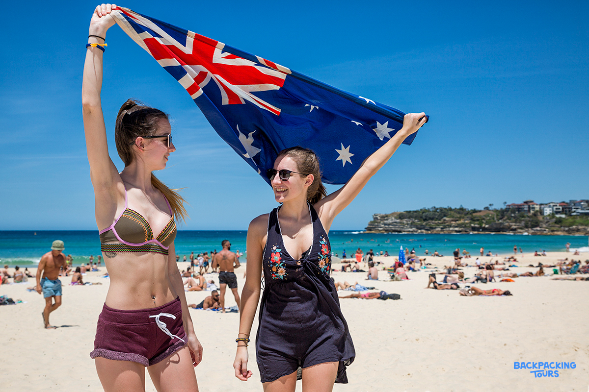 Two joyful women proudly wave the Australian flag on a sunny and crowded Bondi Beach.