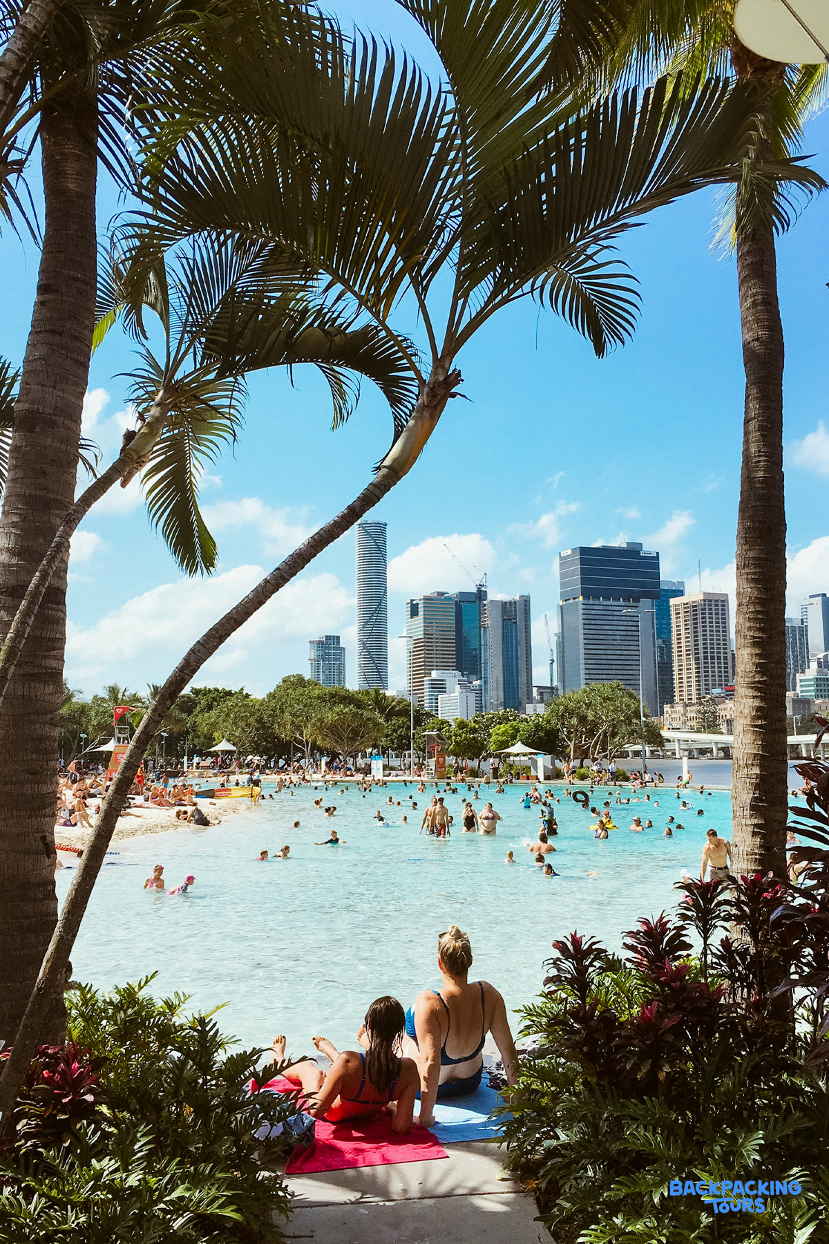 Two women relax by the bustling urban lagoon in Brisbane, framed by palm trees and city skyscrapers.