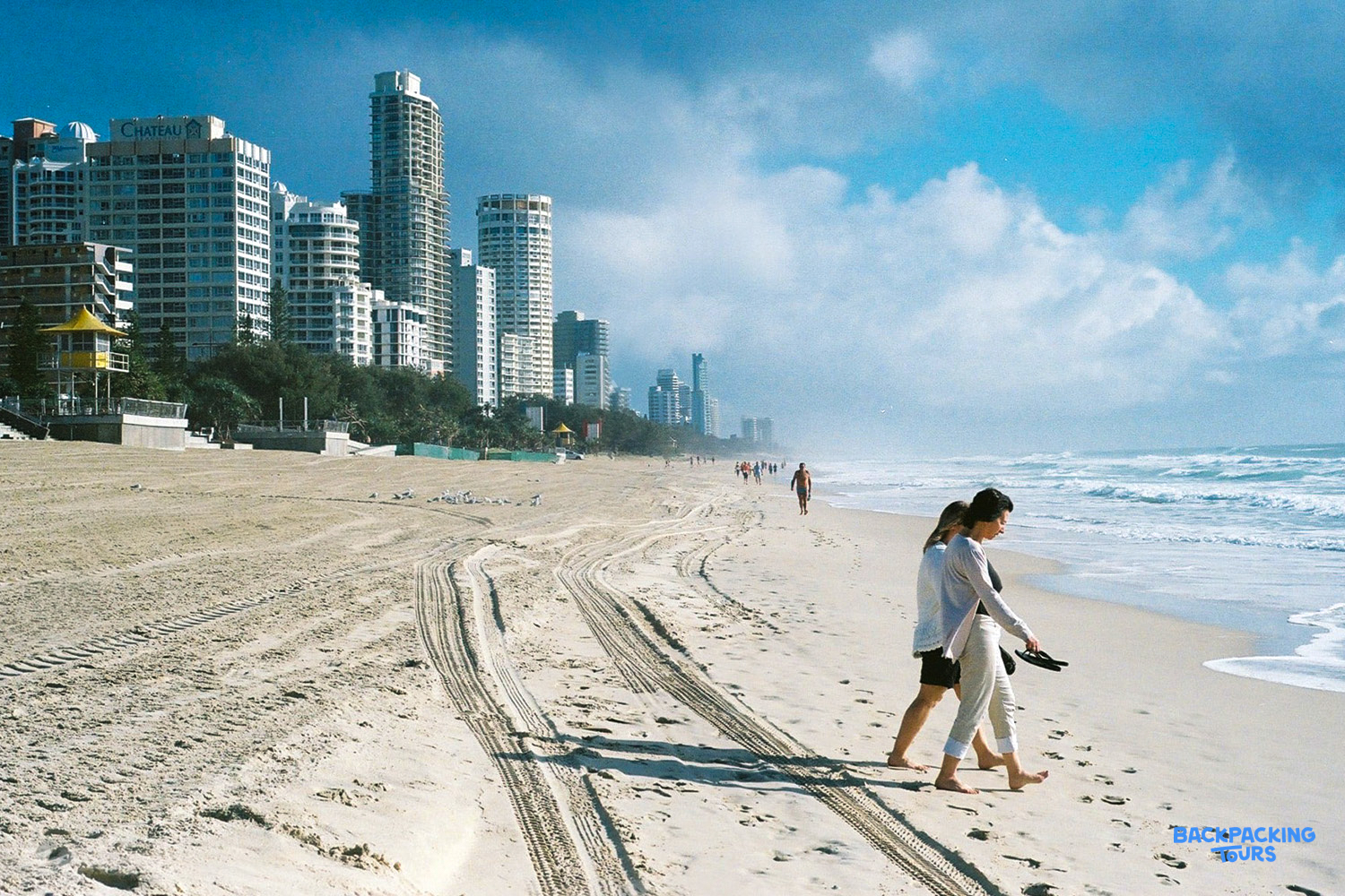 Two people walk barefoot along the golden sands of Surfers Paradise beach, with city skyscrapers in the background.