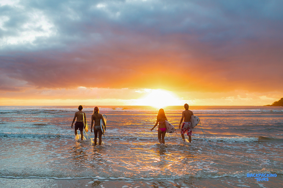 Backpackers carrying surfboards walk into the waves at a vibrant Byron Bay sunset.