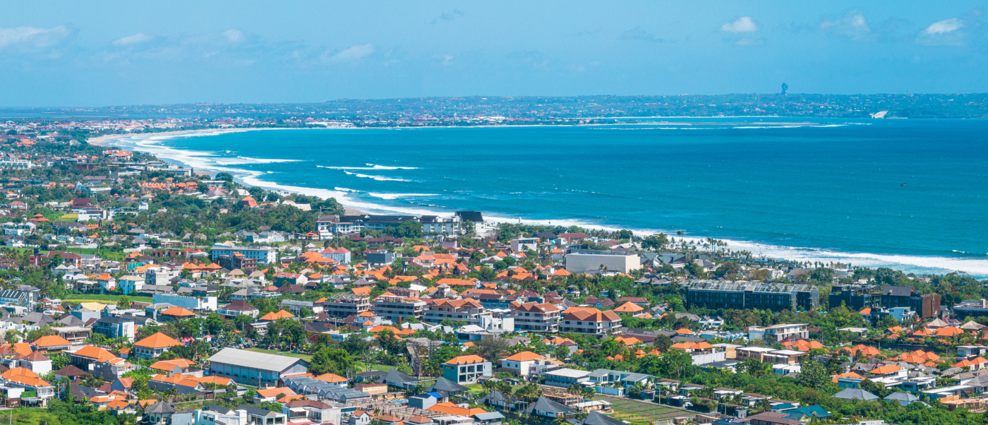 Aerial view of Seminyak, Bali, showing coastal buildings, lush trees, a long beach, and blue ocean.