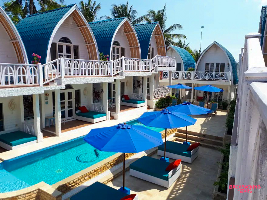 Vibrant blue-roofed bungalows line a sparkling pool with sun loungers at a Gili Trawangan resort.