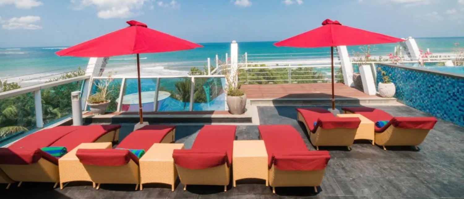 Rooftop terrace with red lounge chairs and umbrellas overlooking the ocean and a pool in Kuta, Bali.