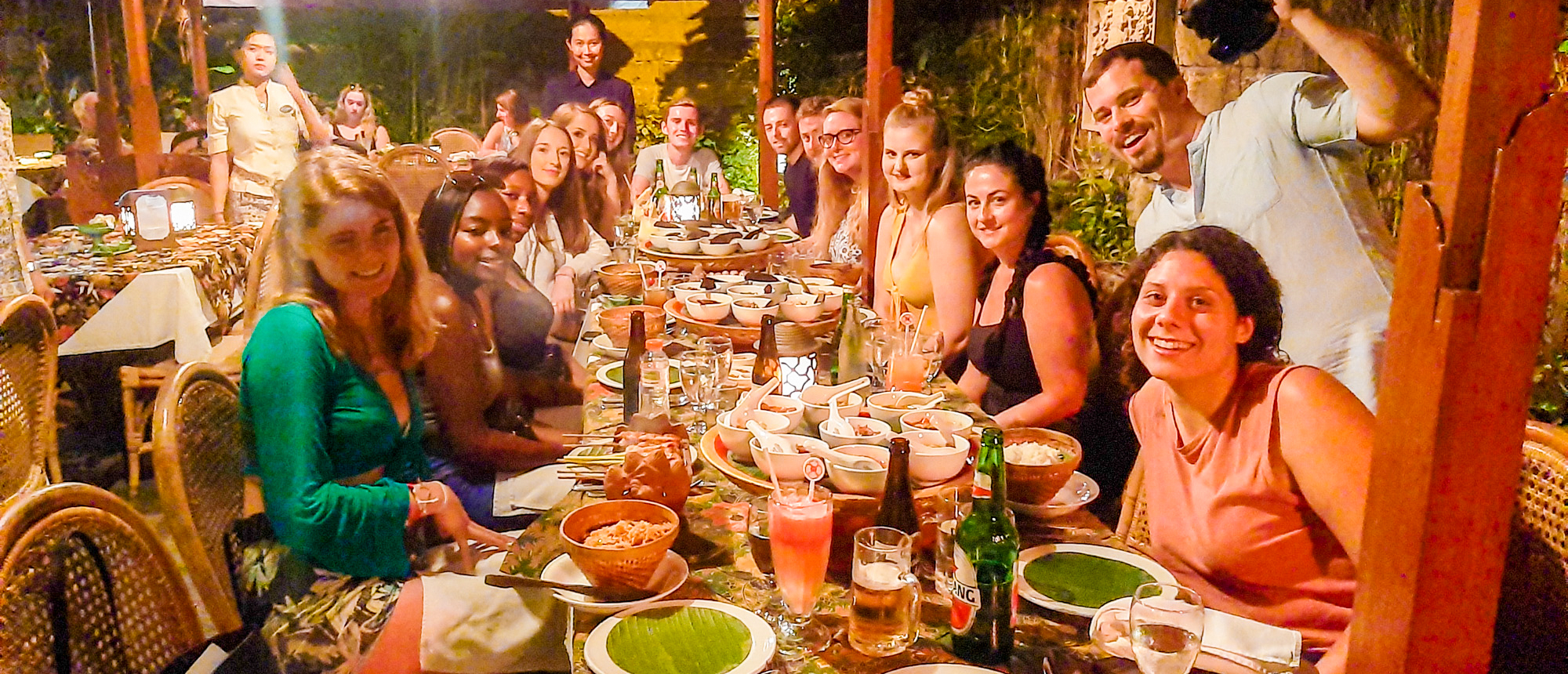 Smiling travelers enjoy a lively welcome dinner with many dishes at an outdoor restaurant in Kuta, Bali.