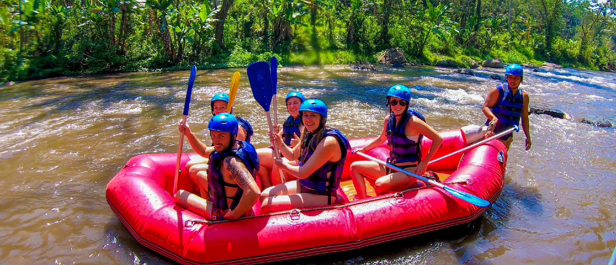 Smiling friends in helmets and life vests raft down a river through dense jungle in Ubud.