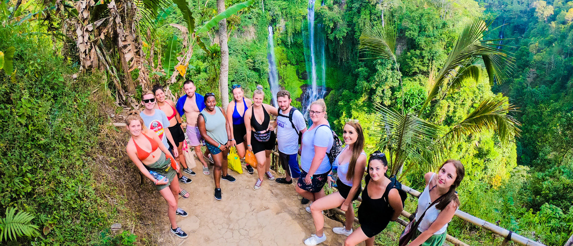 A diverse group of smiling travelers poses on a jungle path overlooking the stunning Sekumpul Waterfalls in Bali.
