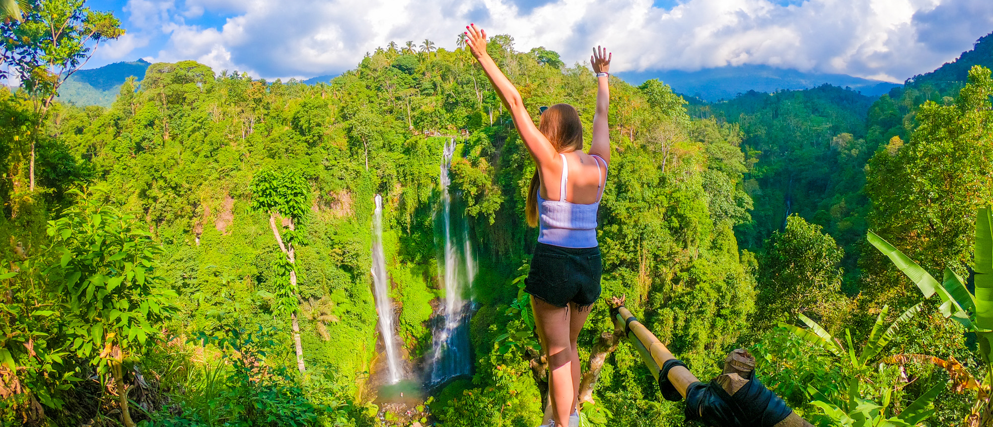 Backpacker with arms raised in joy views the majestic Sekumpul Waterfalls surrounded by dense Balinese jungle.