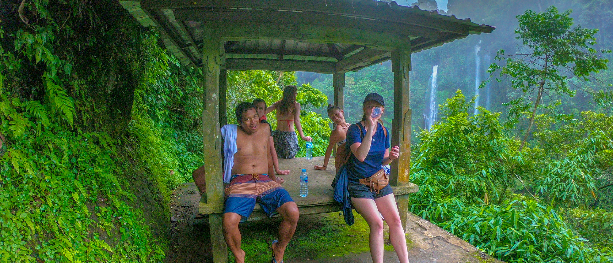 Group of happy backpackers resting in a jungle shelter with stunning Sekumpul Waterfalls in Bali.