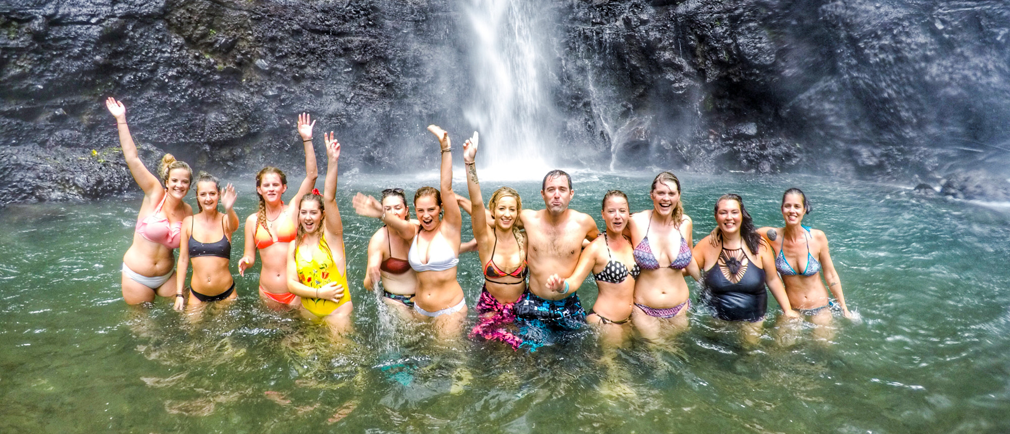 A group of happy travelers waving and swimming in the basin of Sekumpul Waterfall in Bali.