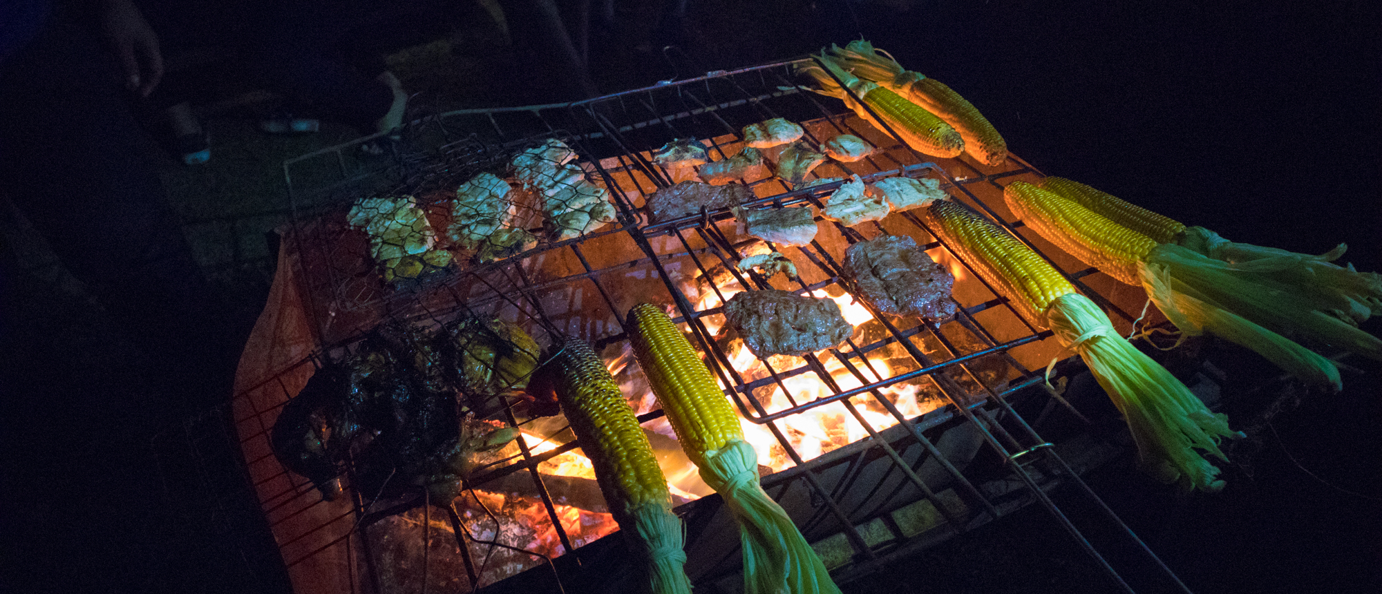 Meat and corn grilling over hot coals at a Bali mountainside camping spot at night.