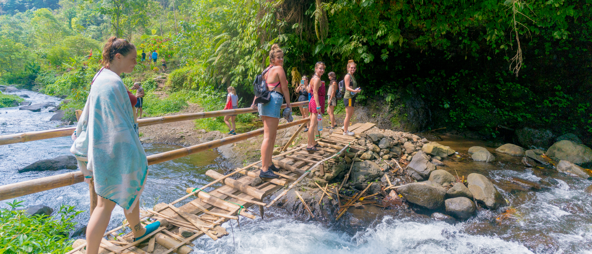 A group of young travelers crosses a rustic bamboo bridge over a river in a lush Bali jungle.