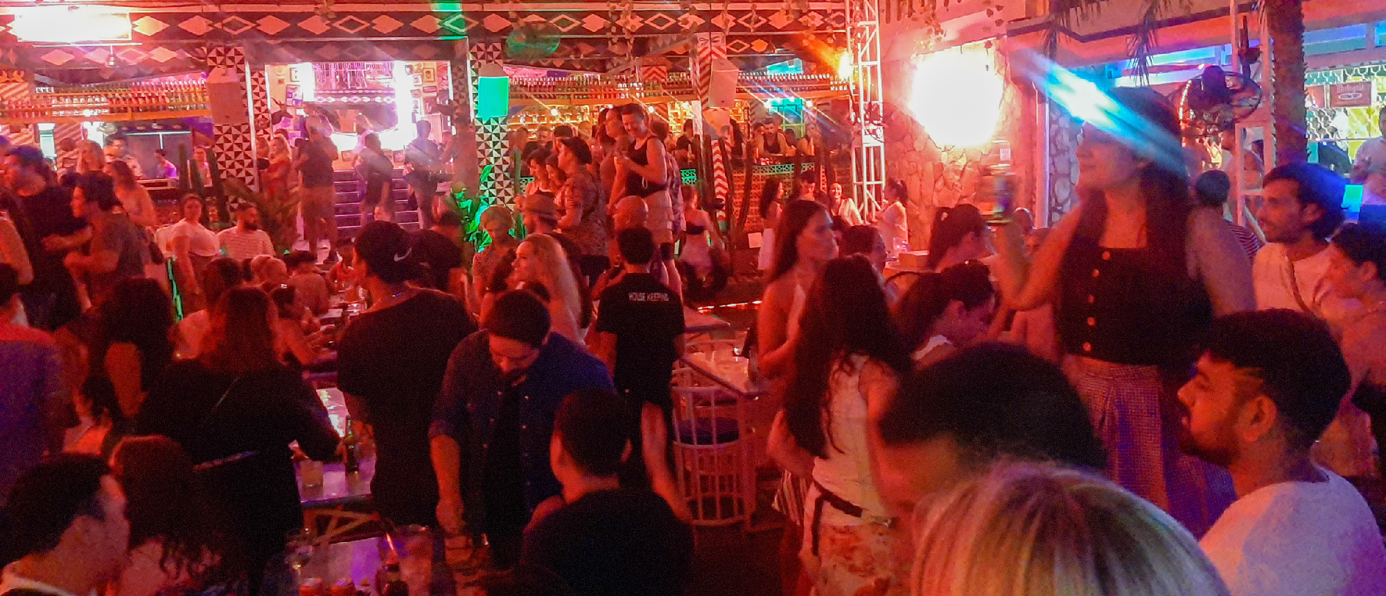 A lively crowd of people dancing and socializing under colorful party lights at a bar in Seminyak.