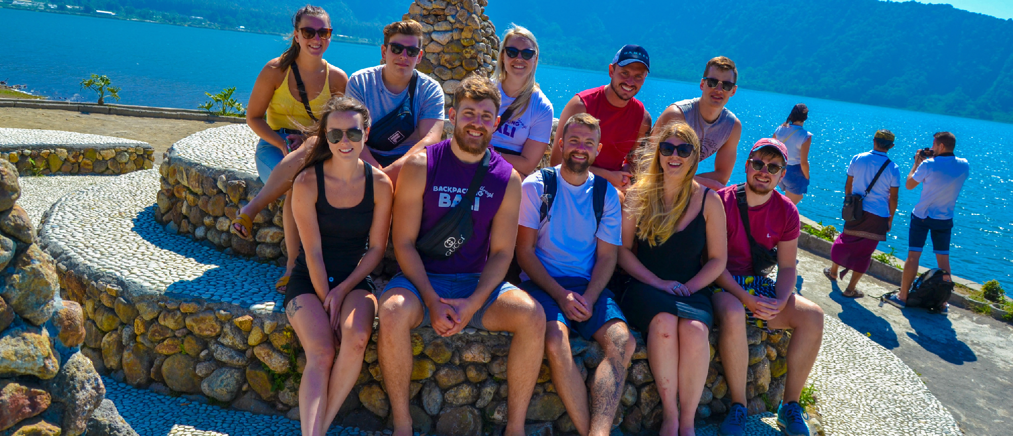 A group of happy backpackers smiling on a stone wall overlooking Lake Beratan in Bali.