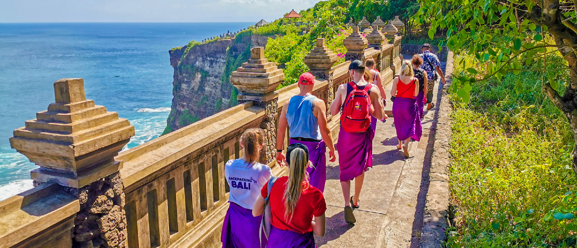 Tourists wearing sarongs walk along a stone path overlooking the ocean at Uluwatu Temple, Bali.