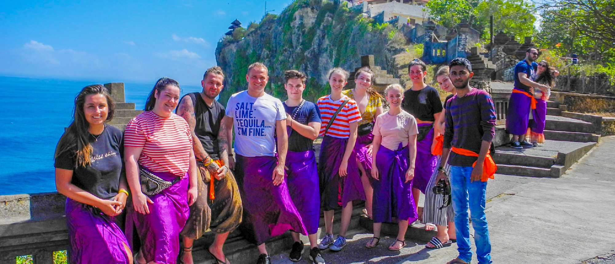 A group of happy backpackers wearing purple sarongs at Uluwatu Temple, Bali, with ocean views.