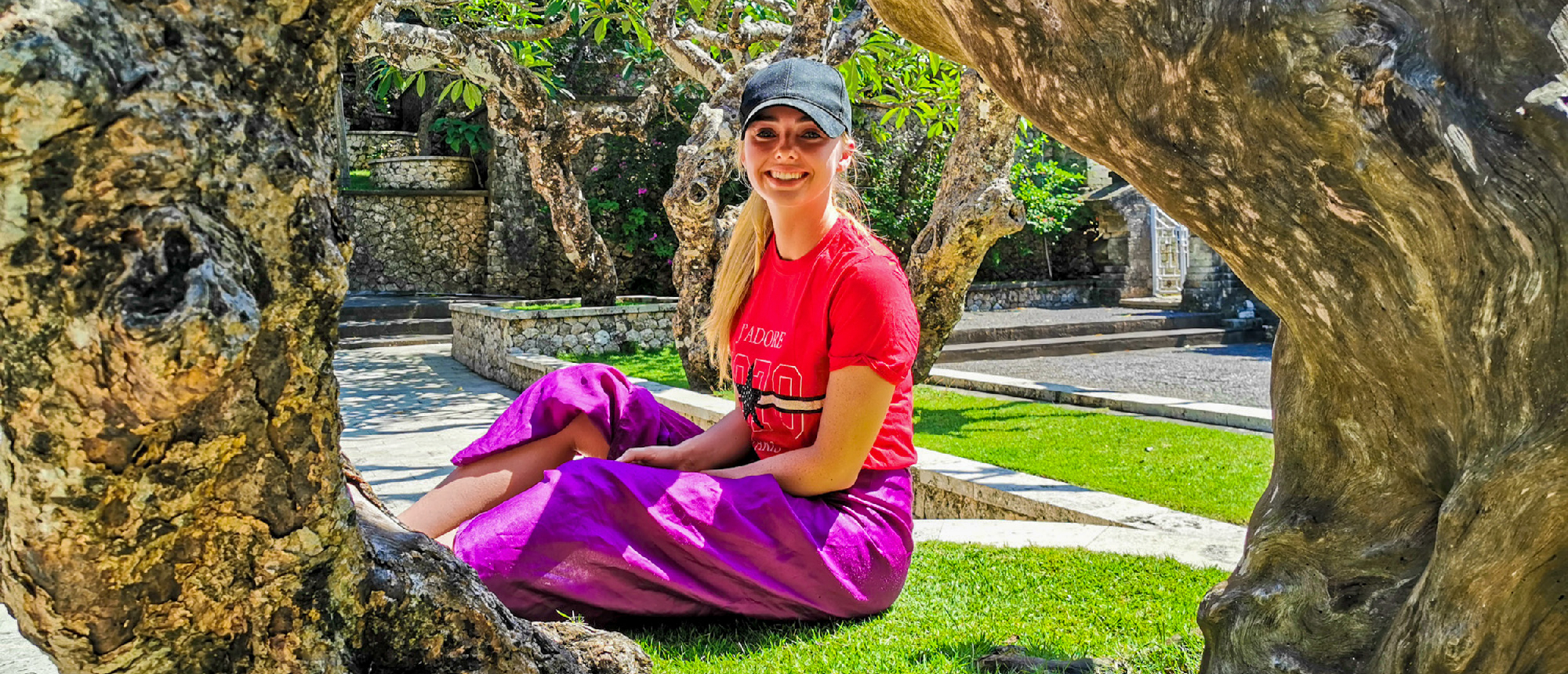 Smiling woman in a purple sarong sits on green grass, framed by gnarled trees in Bali.