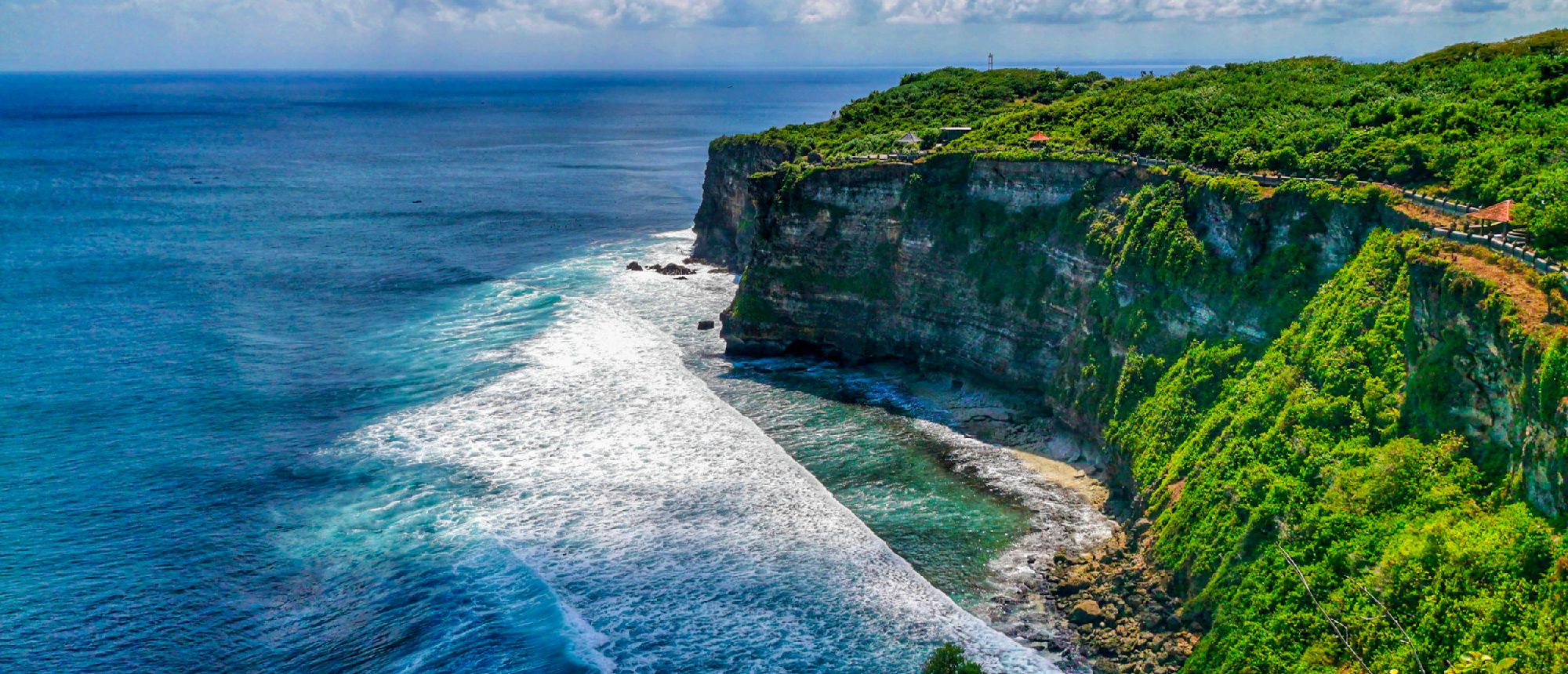 Dramatic green cliffs of Uluwatu, Bali, meet the vibrant blue ocean with white waves crashing.