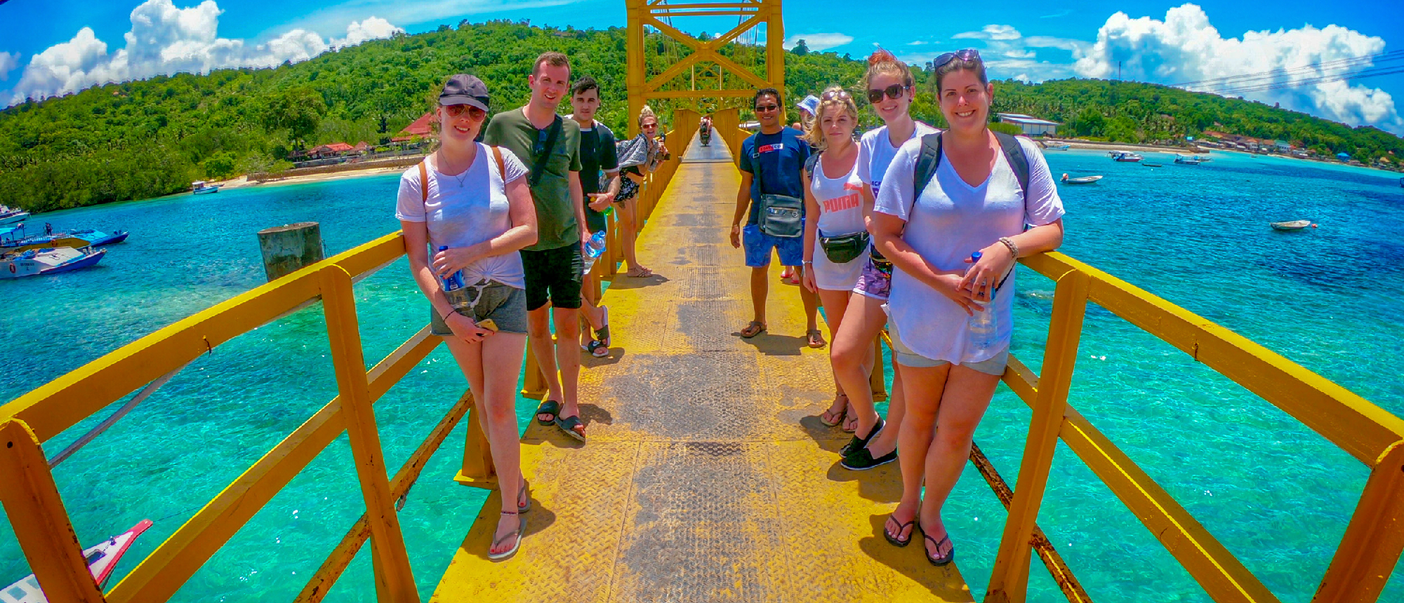 Happy backpackers smile on a bright yellow bridge over clear turquoise water in Nusa Lembongan.