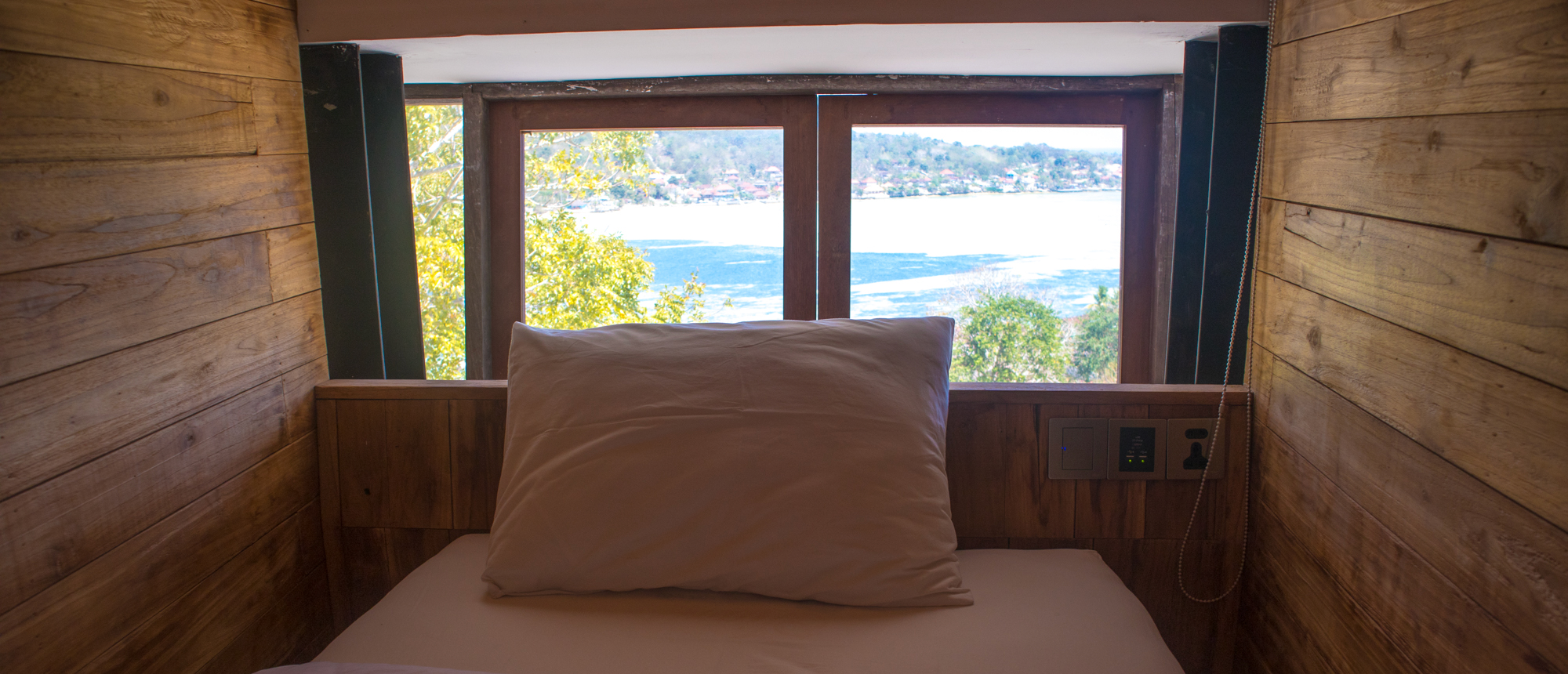 A cozy bed with a white pillow overlooks stunning teal-green ocean waters and distant islands in Nusa Lembongan.