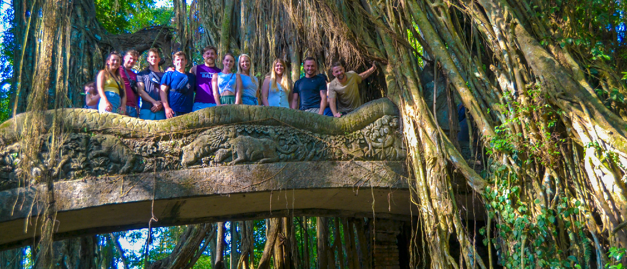A group of backpackers smiling on an ancient stone bridge covered in roots in Ubud's Monkey Forest.