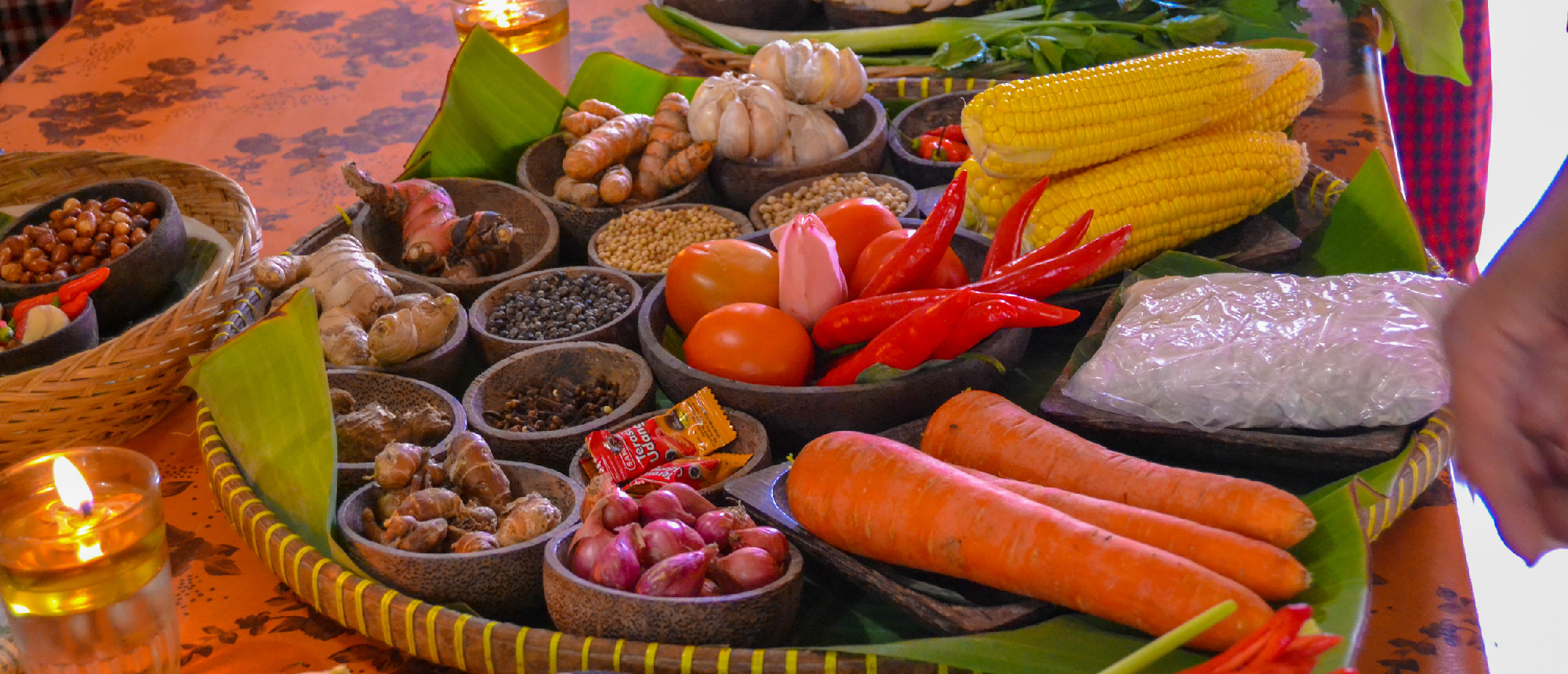 Fresh Balinese cooking ingredients like garlic, chilies, corn, and spices arranged on a woven tray.