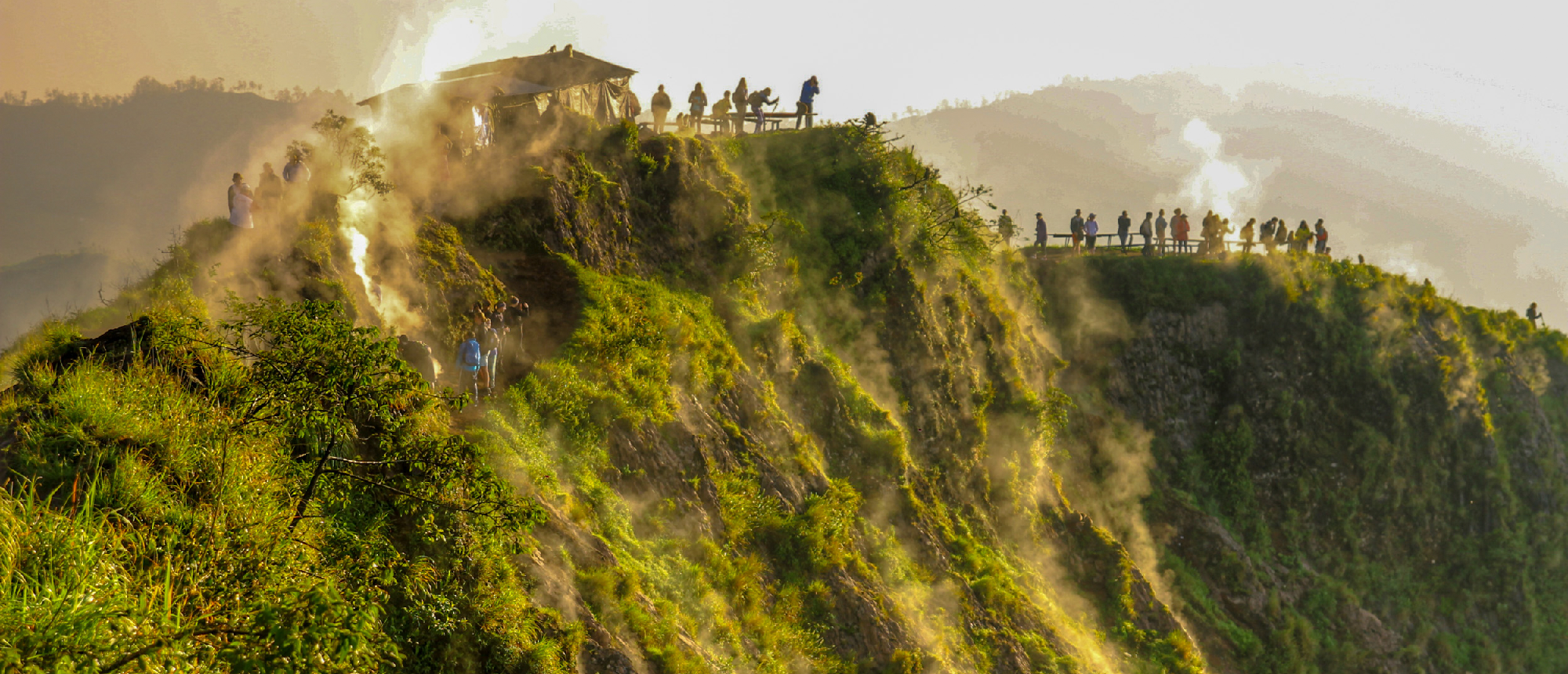Hikers gather on a steaming, green volcanic hillside at sunrise, enjoying the dramatic landscape.