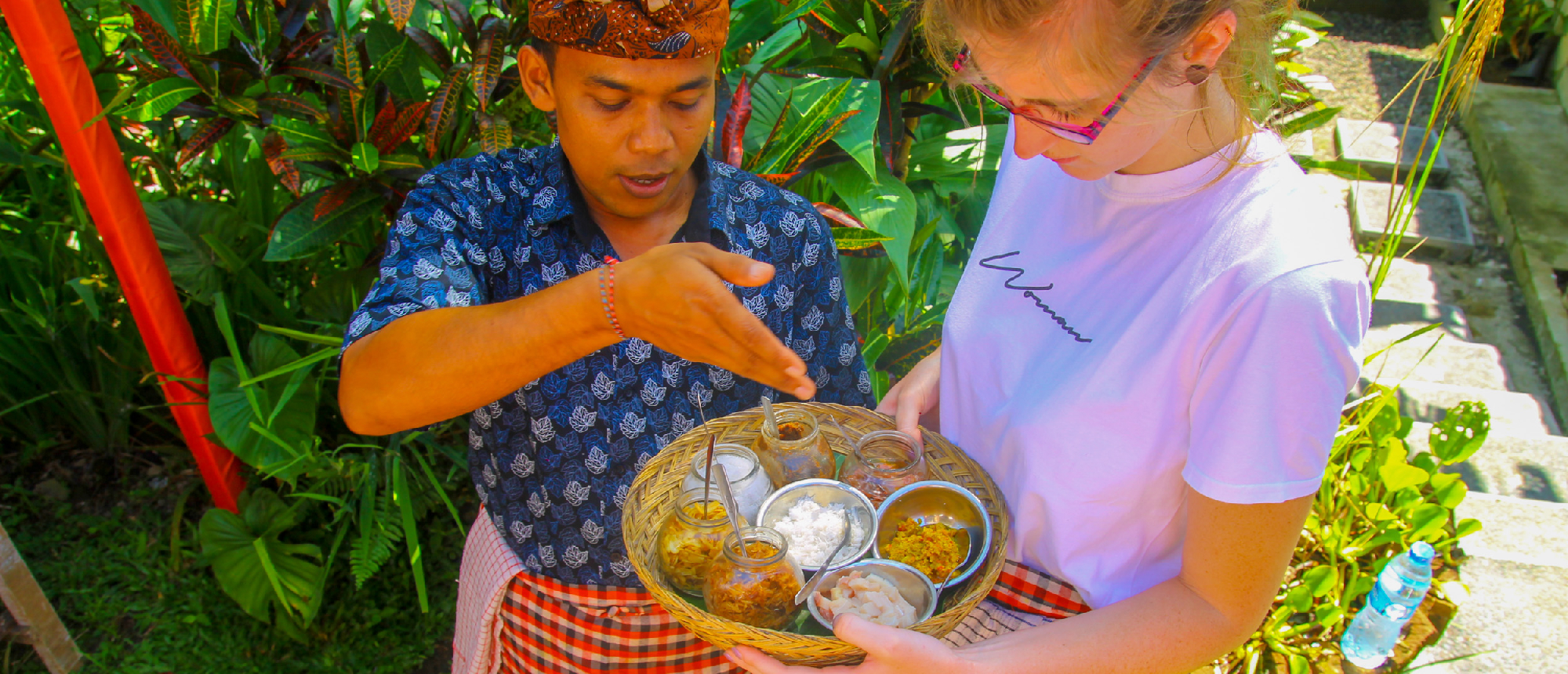 A Balinese man in traditional attire shows a woman ingredients on a woven tray in Ubud, Bali.