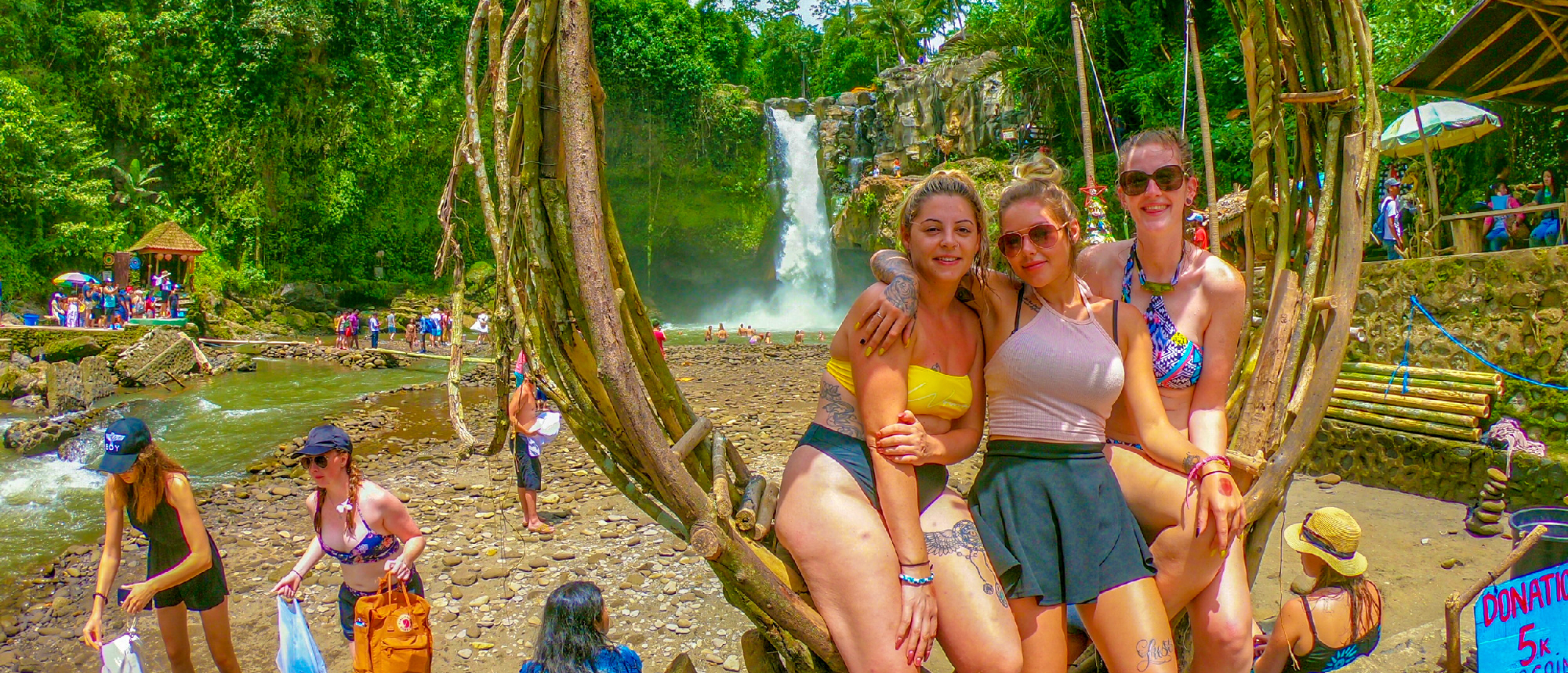 Three smiling women on a unique wooden swing overlooking the majestic Tegenungan Waterfall in Bali.